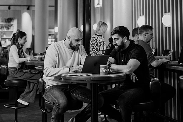 Two members of the Equal Experts Network discuss technology while sat at a laptop in a busy coworking office.
